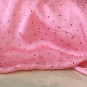 May include: Pale pink fabric with a repeating pattern of tiny, dark grey cross motifs. The fabric is draped, showing gentle folds and shadows. The material appears smooth and delicate, suggesting a lightweight textile.