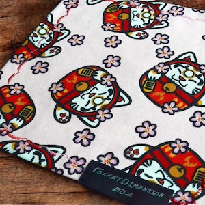 May include: A light-coloured fabric featuring a repeating pattern of red and white beckoning cat figures adorned with small pink cherry blossom flowers. A black rectangular fabric tag is attached to the corner with the text Pocket Dimension EDZ.