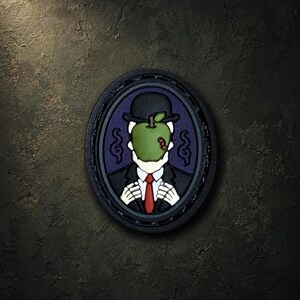 May include: A circular patch featuring a skeletal figure wearing a black suit, white shirt, red tie, and a black bowler hat. A large green apple with a small red worm covers the face. The design is set against a dark purple background.