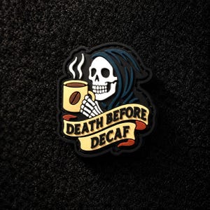 Puede incluir: Un parche de goma que muestra una calavera blanca con capucha azul oscuro, sosteniendo una taza de café amarilla con vapor. Debajo de la calavera, una cinta amarilla con detalles rojos muestra el texto negro DEATH BEFORE DECAF en mayúsculas.