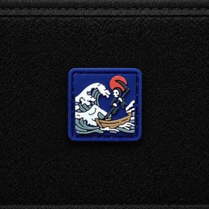 Pode incluir: Um emblema quadrado com borda azul fixado em uma superfície de tecido preto. O emblema apresenta uma ilustração gráfica de um esqueleto em um pequeno barco marrom remando através de uma grande onda branca, com um sol vermelho vivo ao fundo.
