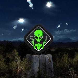Könnte beinhalten: Ein schwarzer und weißer rautenförmiger Patch mit einem grünen Außerirdischen darin. Der Außerirdische macht mit beiden Händen ein Peace-Zeichen. Der Text "51" befindet sich auf beiden Seiten des Außerirdischen. Der Patch befindet sich auf einem Holzpfosten vor einem dunklen, bewölkten Himmel.