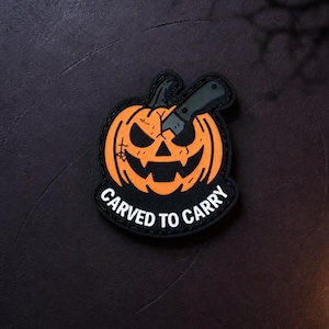 May include: A black and orange morale patch featuring a jack-o'-lantern with a dark green folding knife embedded in its head. The patch has a black border and the white text CARVED TO CARRY printed in a curved arc along the bottom edge.