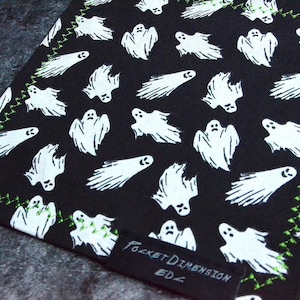 May include: A black fabric featuring a repeating pattern of white ghosts with various expressions. The edges are finished with a bright green zigzag stitch. A small black rectangular tag is sewn onto the corner with the text Pocket Dimension EDZ.