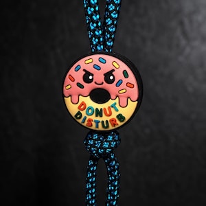 Peut inclure: Un pendentif circulaire représentant un beignet rose avec un visage grincheux, des vermicelles colorés et le texte DONUT DISTURB en relief. Le pendentif est fixé à un cordon tressé noir et bleu qui passe par le centre du motif du beignet.