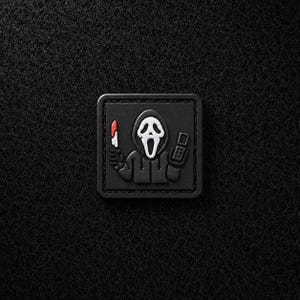 Op de afbeelding: Een vierkante zwarte patch met een verhoogde rubberen afbeelding van een figuur met een capuchon en een wit spookmasker. Het figuur houdt een zilveren mes met een rode bebloede punt in de ene hand en een zwarte mobiele telefoon in de andere.