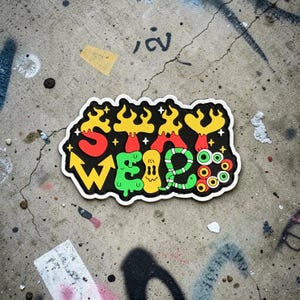 May include: A colorful sticker with the words "STAY WEIRD" in a playful, cartoonish font. The design features red, yellow, green, and black elements with a white border. The sticker is on a concrete surface.