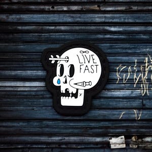 May include: Black and white skull sticker with the text "LIVE FAST" and an infinity symbol. The skull has a single teardrop and a small knife sticking out of its head.