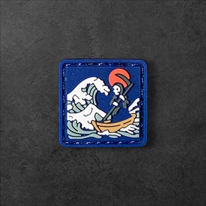 Könnte beinhalten: Ein quadratischer Aufnäher mit blauem Rand zeigt den Sensenmann in einem Boot auf einer Welle, mit einer roten Sonne. Die Welle ist weiß und blau, und das Boot ist hellbraun.