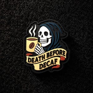 May include: A rubber patch featuring a white skull wearing a dark blue hood, holding a yellow coffee mug with a brown coffee bean graphic and white steam. A yellow banner across the bottom displays the black text DEATH BEFORE DECAF.
