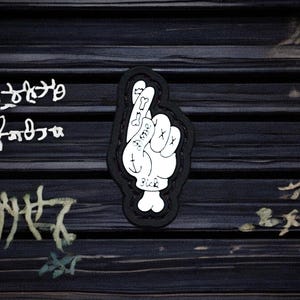 May include: Black and white patch featuring a hand making a 'lucky' gesture. The hand is drawn in a cartoon style with the fingers crossed and the thumb sticking out. The patch has a black border and the words 'Good Luck' are written on the hand.