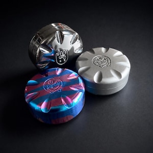 Puede incluir: Tres molinillos metálicos en plata, gris y azul con un patrón de rayas rojas y moradas. Cada molinillo tiene un diseño circular con un emblema estilizado. Los molinillos se muestran sobre una superficie oscura.