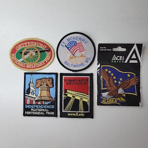 May include: A collection of ten embroidered iron-on patches featuring various American historical sites and landmarks, including Gettysburg National Military Park, Fort McHenry, Independence National Historical Park, the Franklin Institute Science Museum, and Alaska. The patches are all in various colors and feature detailed embroidery.