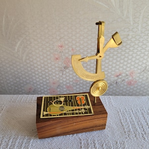 May include: A gold-toned musical scale with a circular base and a decorative wooden stand. The scale features a circular dial with markings and a pointer. The stand has a gold-toned rectangular plate with a design of musical instruments, including a guitar, violin, and flute.