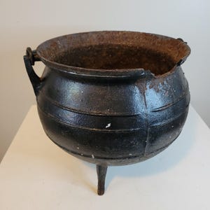 May include: A black cast iron cauldron with three legs and two handles. The cauldron has horizontal bands around its body and a rusty interior. The cauldron is sitting on a white surface.