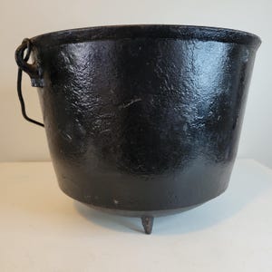 May include: A black cast iron cauldron with a textured surface. The cauldron has a rounded body, a handle on one side, and three small legs. The interior is dark, and the exterior has a glossy finish.