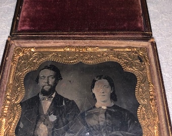 Puede incluir: Una fotografía antigua en tintype de una pareja en un marco dorado. El hombre tiene barba y lleva un traje oscuro. La mujer lleva un vestido oscuro. La fotografía está en una caja forrada de terciopelo.