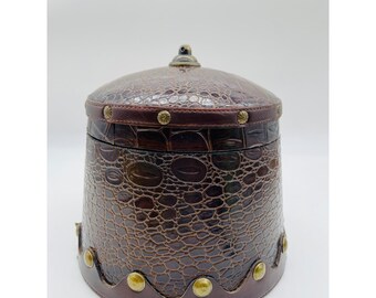 May include: A brown, cylindrical box with a domed lid. The box has a faux crocodile skin texture and is adorned with brass-colored studs. A small, decorative knob sits atop the lid. The box is likely used for storage or decoration.