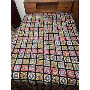 May include: A colorful crocheted blanket with a black background and a geometric pattern of squares and flowers. The blanket is made with a variety of colors, including red, orange, yellow, green, blue, and purple.