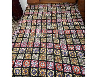 May include: A colorful crocheted blanket with a black background and a geometric pattern of squares and flowers. The blanket is made with a variety of colors, including red, orange, yellow, green, blue, and purple.