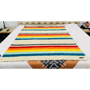 May include: A cream-colored blanket with horizontal stripes in various colors, including red, orange, yellow, blue, and black. The blanket is made of a soft, plush material and is spread out on a bed. A brown tag is sewn on the bottom right corner.