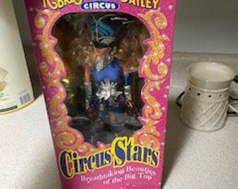 Può includere: Una bambola vintage del circo Ringling Bros. e Barnum & Bailey in una scatola. La bambola indossa un costume blu e argento con un cappello blu e una maschera. La scatola è rosa e bianca con le parole "Ringling Bros. e Barnum & Bailey Circus" e "Circus Stars" stampate su di essa.