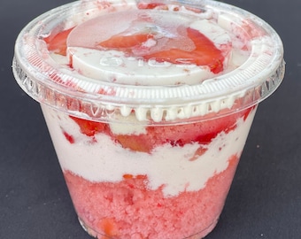 May include: A clear plastic cup filled with layers of pink cake, whipped cream, and fresh strawberries.