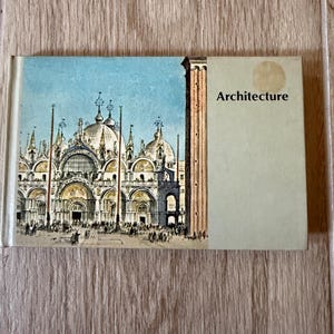 May include: A rectangular hardcover book titled Architecture, featuring a detailed illustration of St. Mark's Basilica in Venice on the left side and a plain light green panel with the word Architecture printed in black on the right side.