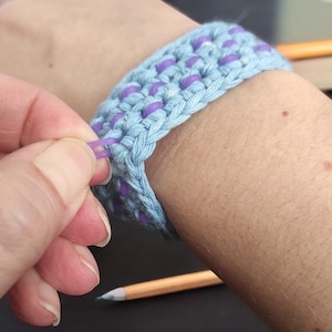 May include: A light blue crocheted bracelet with purple beads. The bracelet is being worn on a person's wrist.