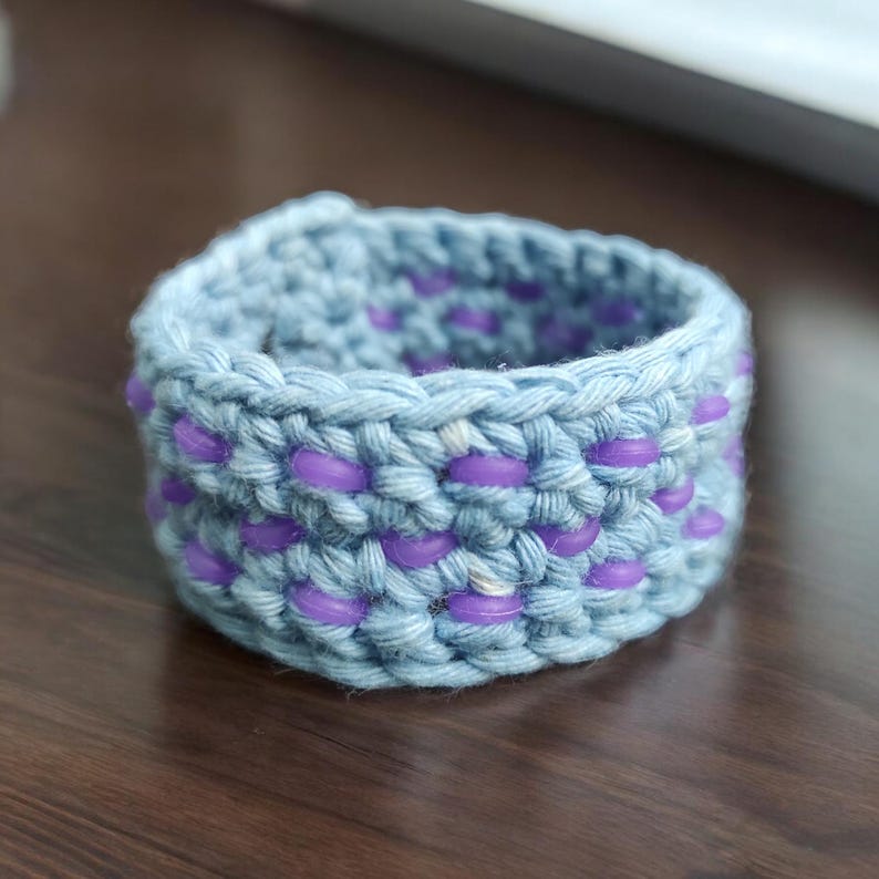 Puede incluir: Un brazalete de crochet azul claro con cuentas moradas tejidas en el dise&ntilde;o.