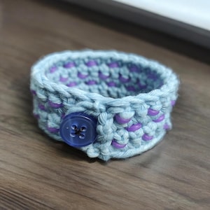 Puede incluir: Un brazalete de crochet azul claro con cuentas moradas y un cierre de bot&oacute;n azul.