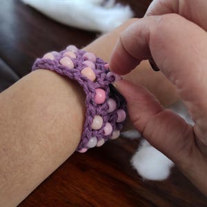 Puede incluir: Un brazalete de crochet morado con cuentas rosas. El brazalete se está abrochando con un cierre.