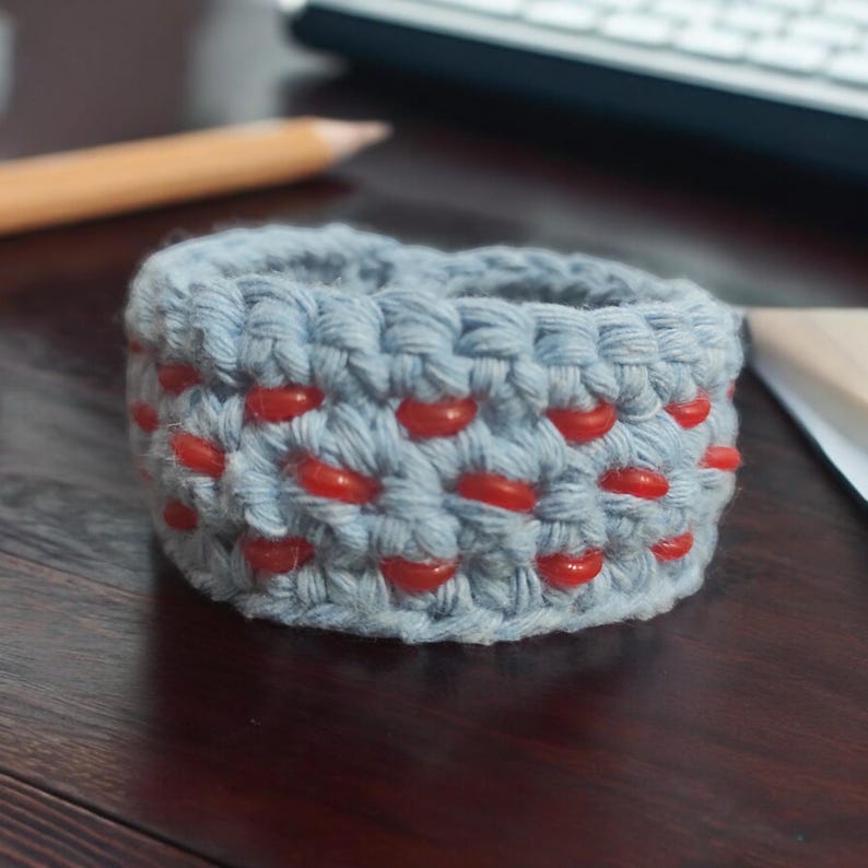 Puede incluir: Un brazalete de crochet azul claro con cuentas rojas. El brazalete est&aacute; hecho con un patr&oacute;n de punto simple y tiene un brillo sutil.