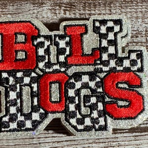 May include: Embroidered patch with the word "BULLDOGS" in red and black checkered letters. The letters are outlined in silver and have a glittery effect. The patch is likely for a sports team or school.