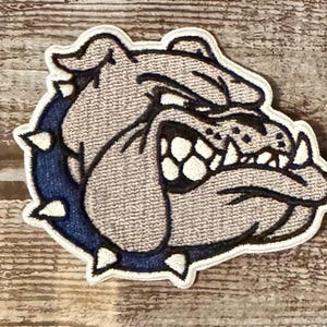 May include: Embroidered patch featuring a bulldog's head with a fierce expression. The patch has a light gray background with a blue collar and white accents. The bulldog's teeth are prominently displayed, and the patch is outlined in white.