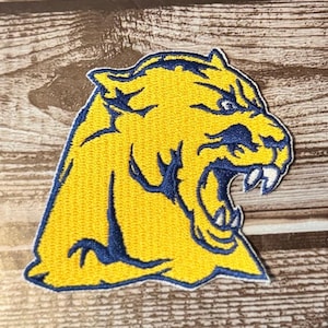 May include: Embroidered patch featuring a snarling cougar head. The cougar is yellow with navy blue accents, including the outline, facial details, and teeth. The patch is likely for a sports team or school.