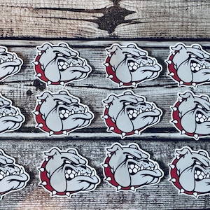 May include: A collection of bulldog head stickers. Each sticker features a gray bulldog head with a red spiked collar. The stickers are arranged on a wooden surface.