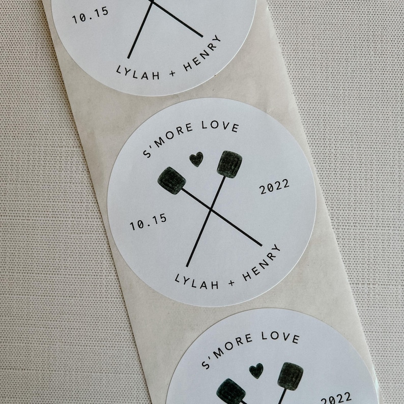 Puede incluir: Pegatina redonda blanca con texto negro y un gr&aacute;fico de dos palos cruzados con malvaviscos. El texto dice "S'more Love", "10.15", "2022", "Lylah + Henry".