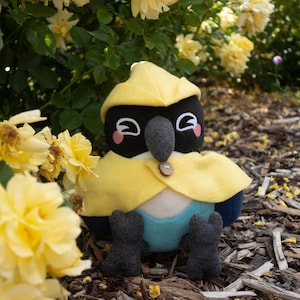 May include: A plush bird toy featuring a black head, a dark grey beak, and pink cheek patches. It wears a yellow hood and cape fastened with a small wooden button, paired with a teal body and dark grey feet, sitting among yellow flowers.