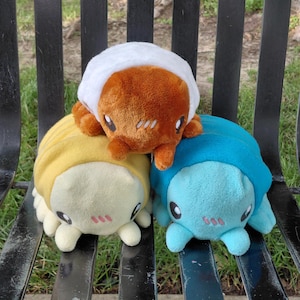 May include: Three plush toys shaped like pill bugs or isopods. One is orange with a white top, one is pale yellow, and one is bright blue. Each plush features large dark eyes and three small pink embroidered lines on the cheeks for detail.