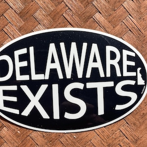 Peut inclure: Un autocollant ovale noir et blanc avec le texte "DELAWARE EXISTS" en lettres blanches. L'état du Delaware est délimité en blanc dans le coin inférieur droit de l'autocollant.