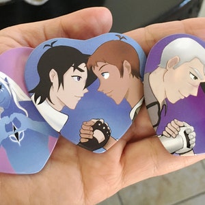May include: Four heart-shaped pins with cartoon characters. The first pin features a blue character with horns and white hair making a heart shape with their hands. The second pin features two characters with black and brown hair looking at each other. The third pin features two characters with white and black hair looking at each other. The fourth pin features two characters with white and black hair looking at each other.