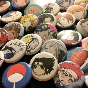May include: A collection of round, colourful anime character buttons. The buttons feature a variety of characters, including a girl with pink hair, a boy with black hair, and a character with a red scarf. The buttons are all approximately 5 cm in diameter.