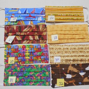 May include: Six different fabric face masks with numbers 25 through 32 written on yellow sticky notes. The masks are all pleated and have different patterns, including sailboats, feathers, puzzle pieces, flowers, and cowboys.