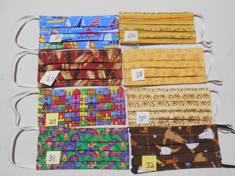 May include: Six different fabric face masks with numbers 25 through 32 written on yellow sticky notes. The masks are all pleated and have different patterns, including sailboats, feathers, puzzle pieces, flowers, and cowboys.