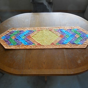 May include: A colorful table runner with a geometric pattern in shades of orange, yellow, green, blue, and purple. The runner is made of fabric and has a quilted texture.