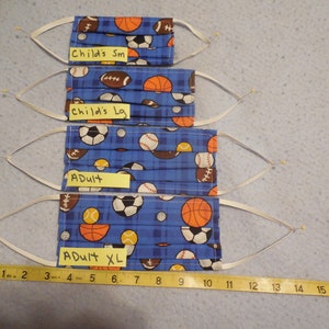 May include: Four pleated face masks with elastic ear loops. The masks are blue with a sports-themed pattern of footballs, basketballs, soccer balls, and baseballs. Sizes include Child's Sm, Child's Lg, Adult, and Adult XL. A measuring tape is included.