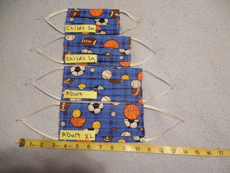 May include: Four pleated face masks with elastic ear loops. The masks are blue with a sports-themed pattern of footballs, basketballs, soccer balls, and baseballs. Sizes include Child's Sm, Child's Lg, Adult, and Adult XL. A measuring tape is included.
