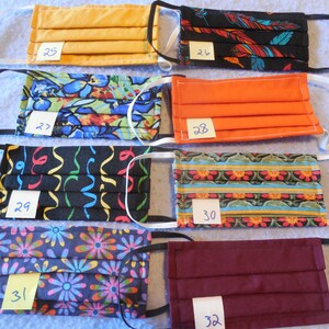 May include: A collection of eight pleated face masks in various colors and patterns. Designs include solid orange, floral, and abstract patterns. Each mask has elastic ear loops and a small tag with a number.