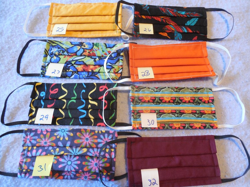 May include: A collection of eight pleated face masks in various colors and patterns. Designs include solid orange, floral, and abstract patterns. Each mask has elastic ear loops and a small tag with a number.